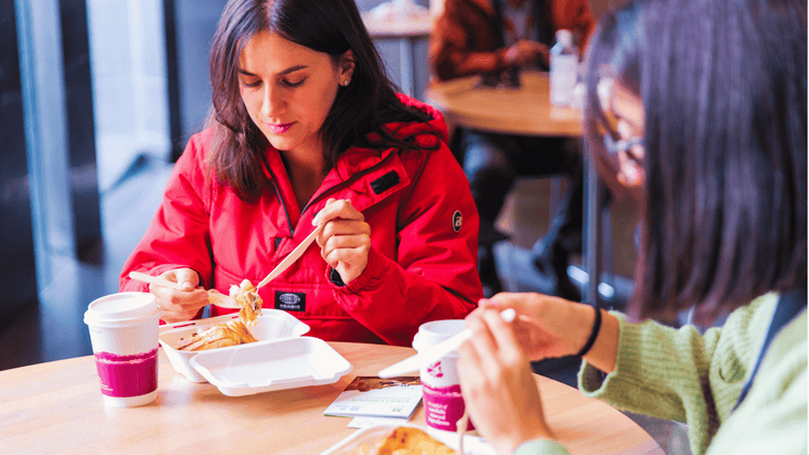 2 visitors enjoying a meal at a table in the Giant's Causeway Visitor Centre Café, with takeaway coffee cups and food containers in a bright indoor seating area and other café guests sitting in the background